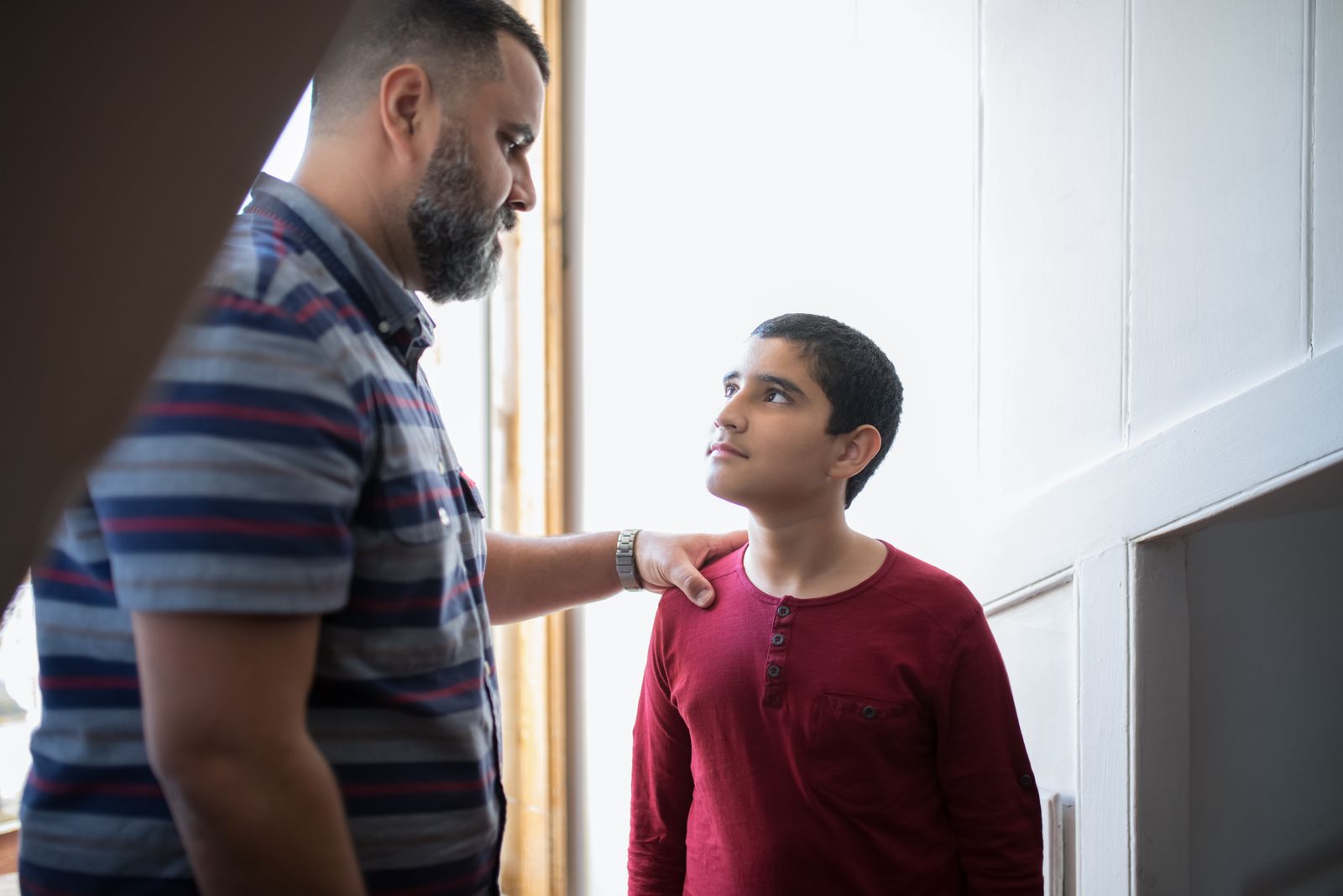A father sitting at a table across from his young son, both looking down at the boy's homework. The son appears thoughtful rather than fearful. In the background, a calendar on the wall and a bookshelf suggest a home where consistency and teaching are valued. The image captures a moment of correction as relationship, not punishment—a father's calm presence and attention focused on helping his child think through what he did and why it matters.