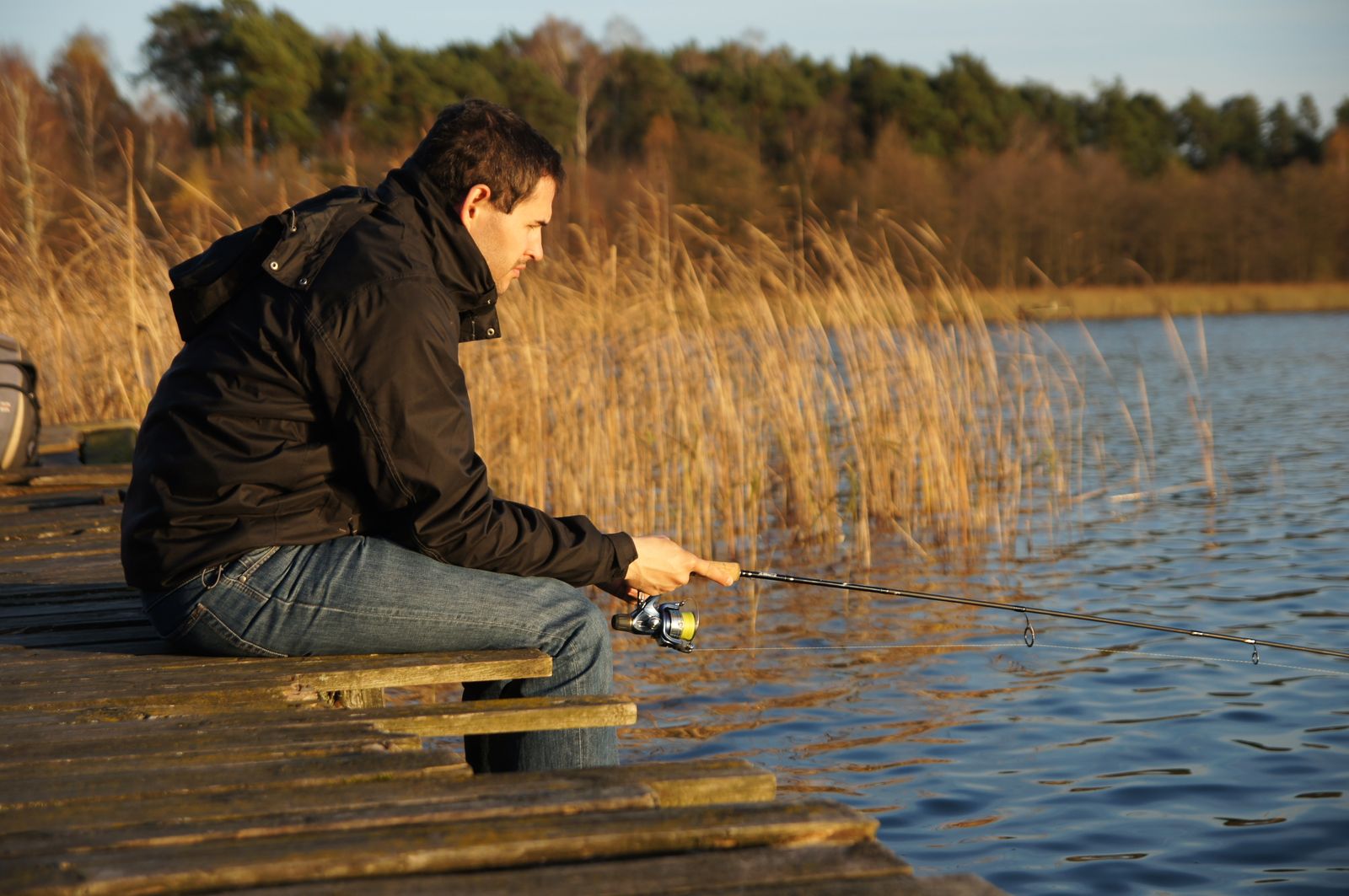 A man in a baseball cap and waders stands on the shore of a calm lake, casting a spinning rod toward the water. His tackle box rests on a nearby rock, and pine trees line the distant bank. The morning light is soft and flat, suggesting early dawn.