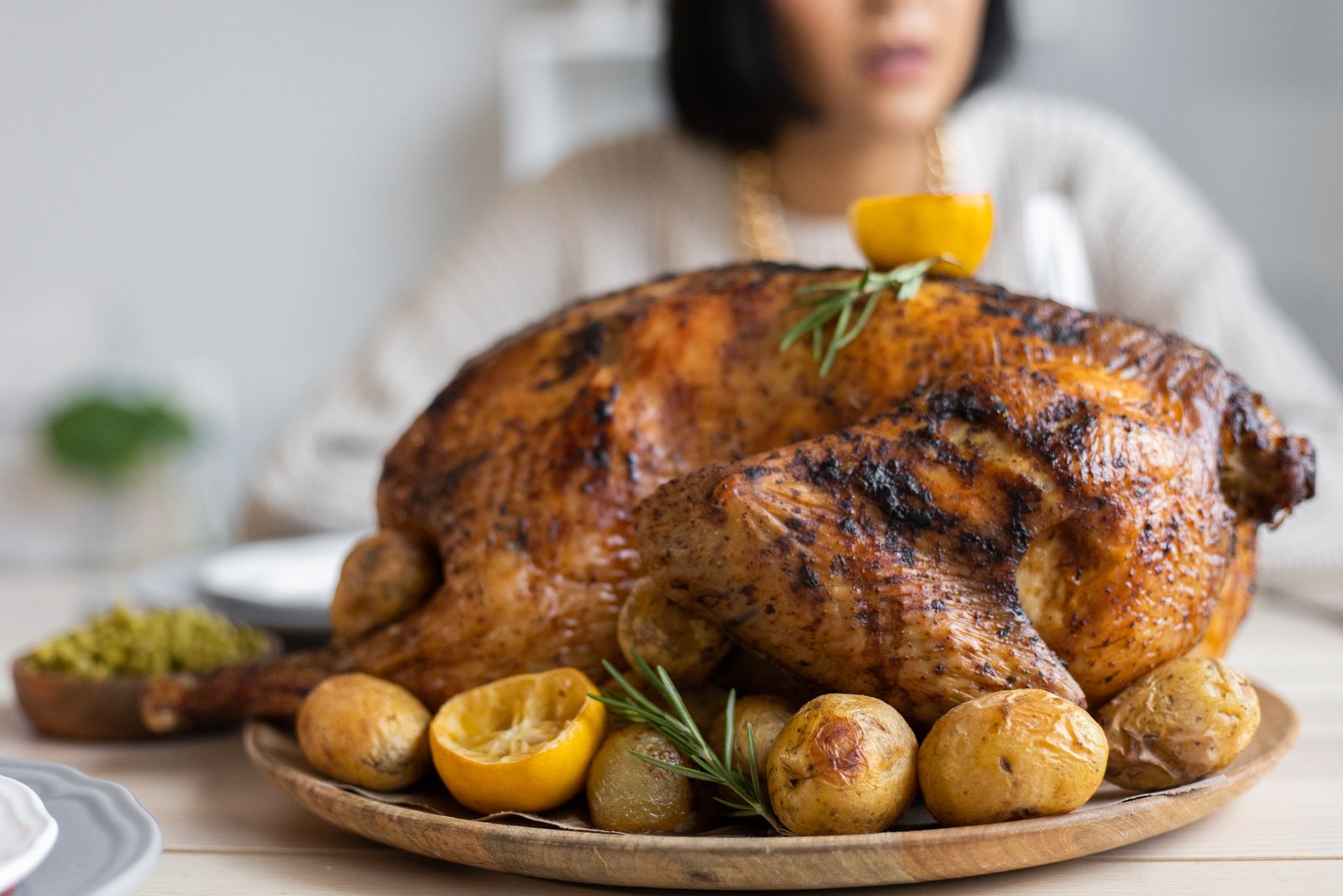 A chef in an overhead view using a sharp knife to carve a golden roasted turkey on a wooden cutting board, with a carving fork steadying the bird and the first thigh being removed. A meat thermometer is visible inserted into the thigh, and a warm serving platter waits nearby.