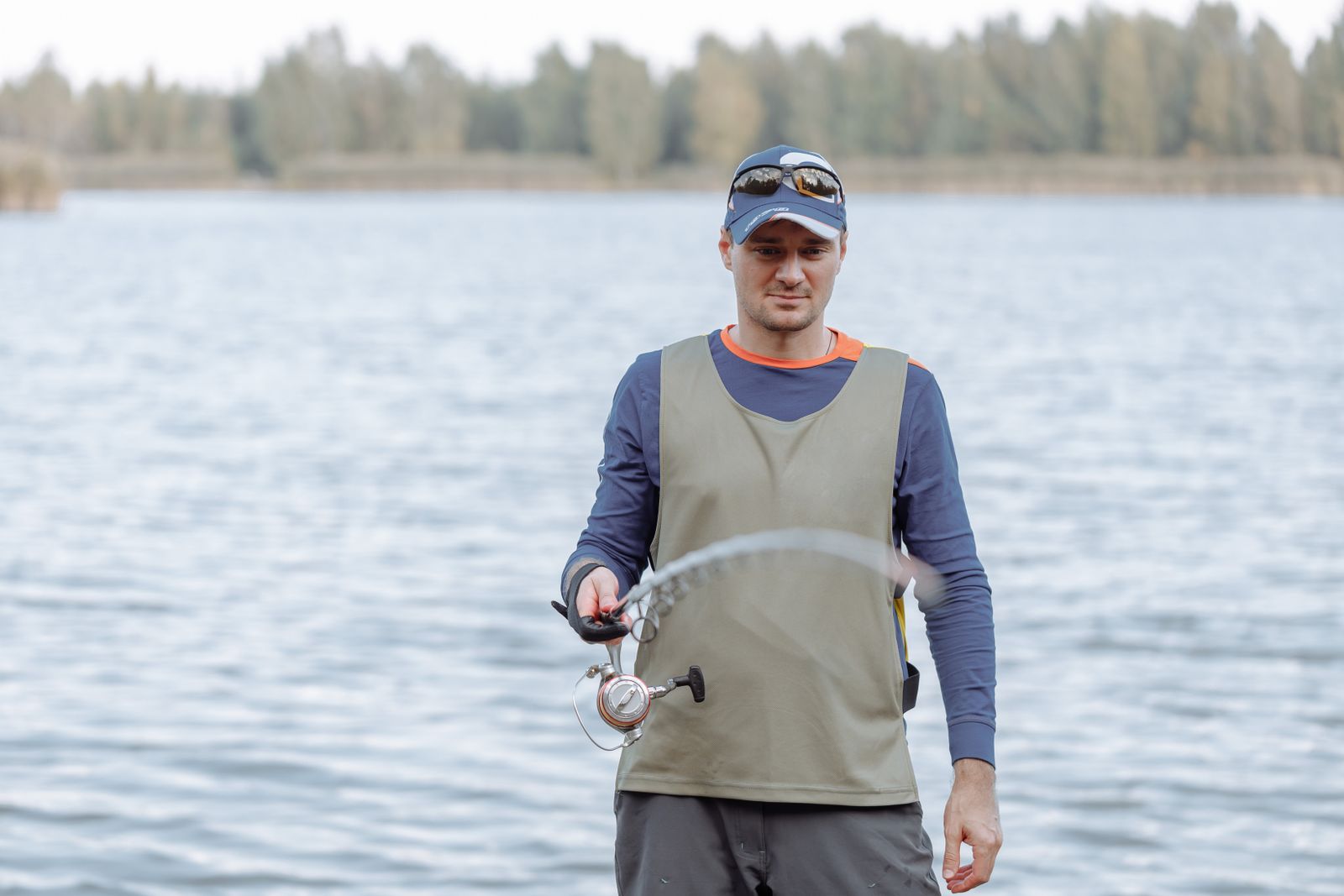 Close-up of a man's hand positioned on a baitcasting reel, thumb resting on the spool with firm contact, rod angled backward at one o'clock position, showing the grip and thumb pressure required during the backcast phase.