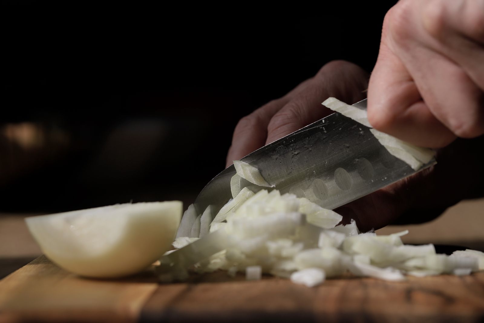 Overhead diagram showing a halved onion positioned root-end-down on a cutting board, with dotted lines indicating horizontal cuts through the onion layers, followed by vertical parallel cuts perpendicular to the root, and a second set showing crosswise cuts creating uniform dice. Inset shows a left hand in proper claw grip position with knuckles forward and fingertips curled under, demonstrating the guide-hand position relative to the blade.