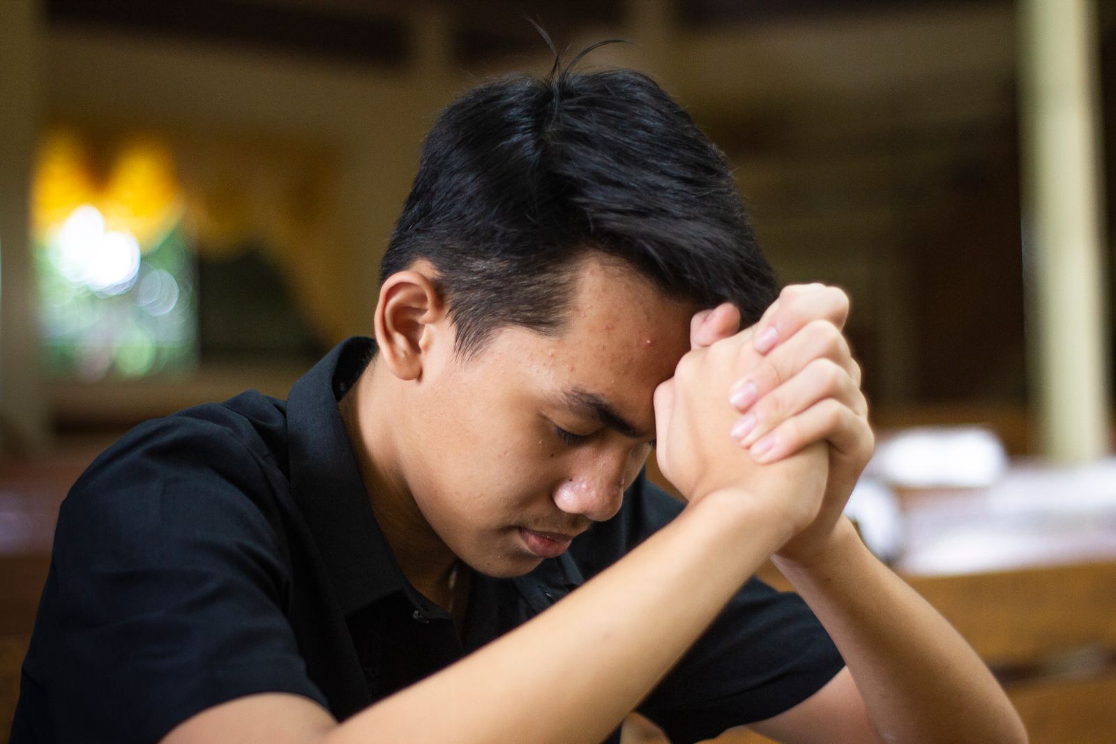 A man sitting alone in early morning light with his head bowed and hands folded, an open Bible beside him on the table.