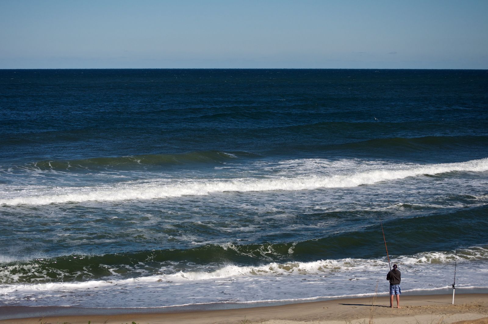 A person standing waist-deep in Atlantic Ocean surf at dawn, casting a 10-foot surf rod toward a visible sandbar with breaking waves, holding a sand spike rod holder in their other hand, with labeled beach structures showing the trough between shore and sandbar, and a cut through the sandbar where no waves break.</illustrationAlt>
</invoke>